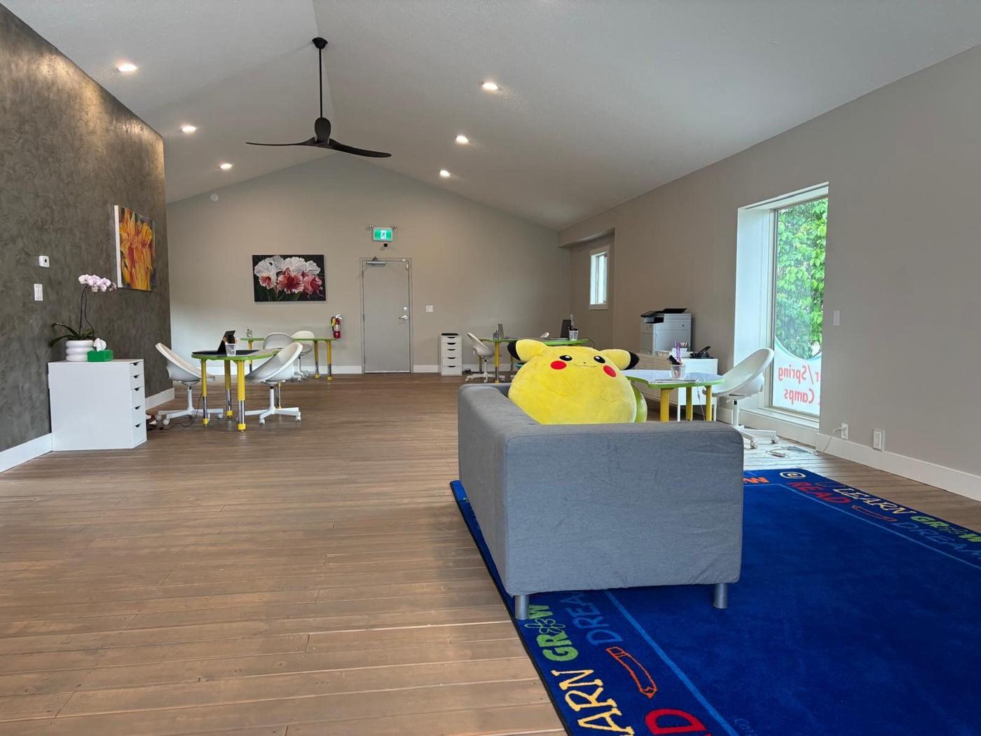 A bright main room with wood floors, small tables and chairs, a grey couch with a plush toy, and a blue rug