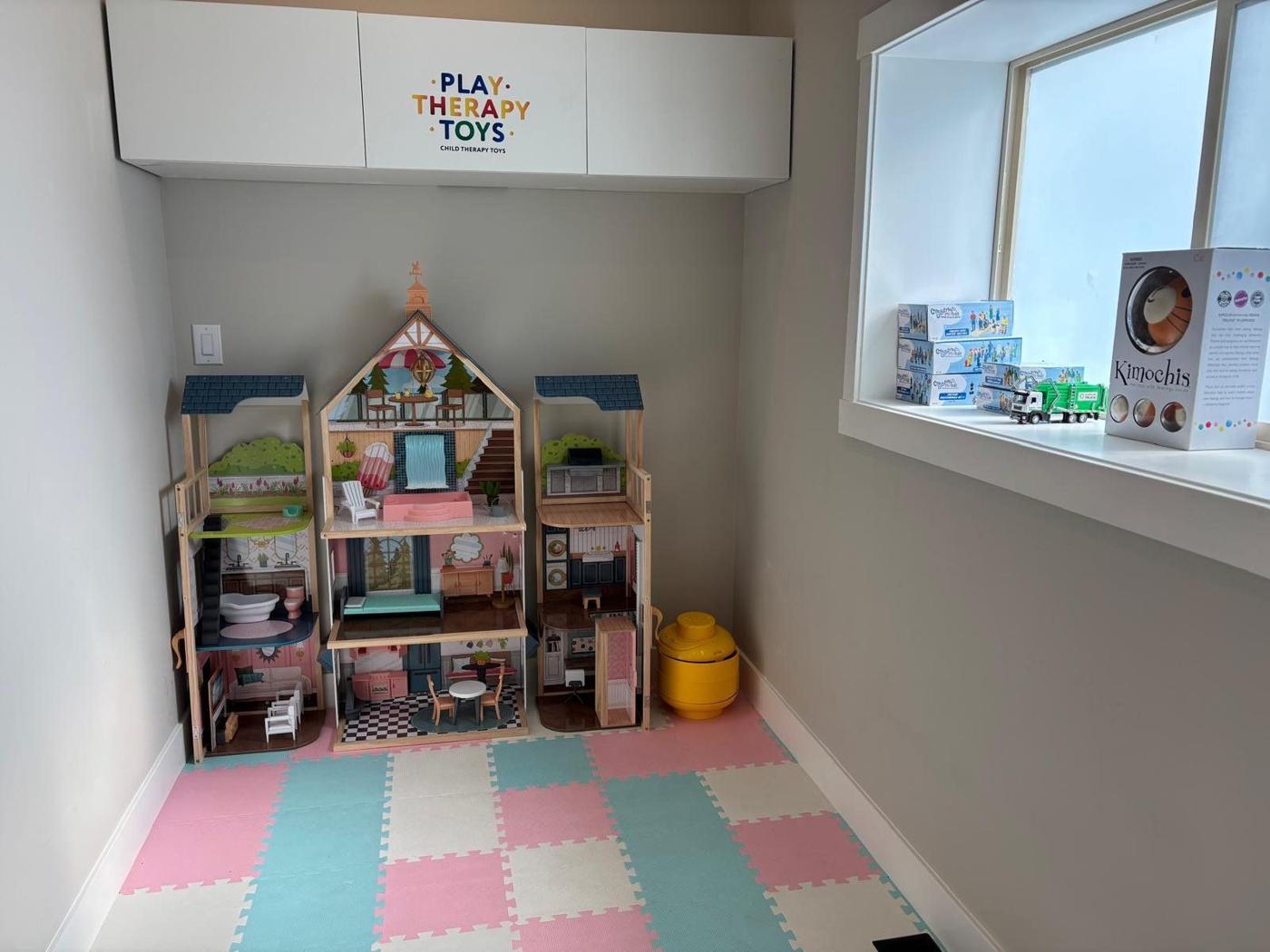 A play room with dollhouses, play therapy toys and soft pink, teal and cream floor mats